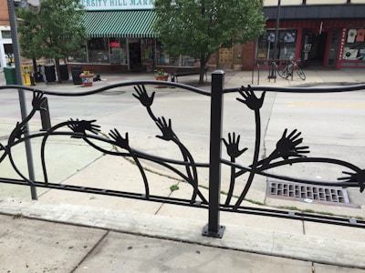 A decorative metal fence features silhouettes of hands reaching outwards in various directions. Behind the fence, there is a street scene with trees, a bicycle, and shops including one with a green and white striped awning labeled 'Hill Market'. The environment is urban and appears calm.