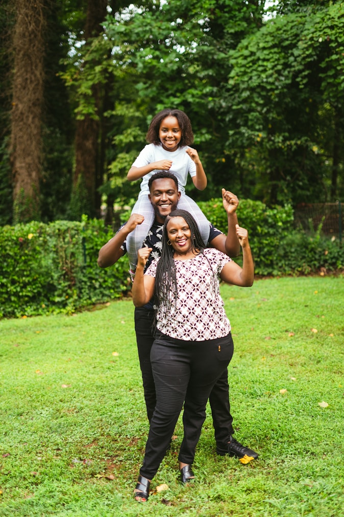 A joyful family scene in a lush green garden. Three individuals are stacked in a playful manner, with a child sitting on the shoulders of a man, who is standing behind a woman. All are smiling and displaying a sense of happiness, surrounded by vibrant greenery.