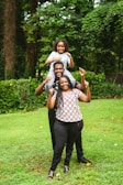 A joyful family scene in a lush green garden. Three individuals are stacked in a playful manner, with a child sitting on the shoulders of a man, who is standing behind a woman. All are smiling and displaying a sense of happiness, surrounded by vibrant greenery.