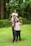 A joyful family scene in a lush green garden. Three individuals are stacked in a playful manner, with a child sitting on the shoulders of a man, who is standing behind a woman. All are smiling and displaying a sense of happiness, surrounded by vibrant greenery.
