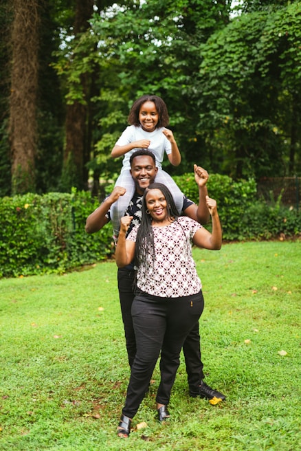 A joyful family scene in a lush green garden. Three individuals are stacked in a playful manner, with a child sitting on the shoulders of a man, who is standing behind a woman. All are smiling and displaying a sense of happiness, surrounded by vibrant greenery.
