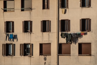 A beige apartment building facade with several windows that have dark brown shutters. Some windows are open, and there are various pieces of clothing and laundry hanging on clotheslines strung between the windows.