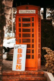 An orange telephone booth is situated on a brick sidewalk. In front of it, there is a sign that reads 'Come in we are open' with bold orange letters. The area around the booth features some greenery and outdoor elements.