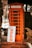 An orange telephone booth is situated on a brick sidewalk. In front of it, there is a sign that reads 'Come in we are open' with bold orange letters. The area around the booth features some greenery and outdoor elements.