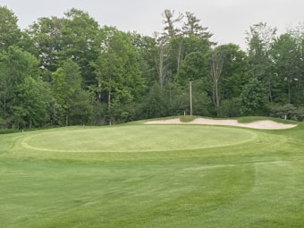 A lush golf course with a well-maintained green and sand bunkers. Surrounding the course are dense trees and foliage.
