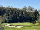 A lush green golf course featuring well-manicured grass and several sand bunkers is surrounded by dense, leafy trees. The clear blue sky enhances the picturesque landscape.