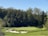 A lush green golf course featuring well-manicured grass and several sand bunkers is surrounded by dense, leafy trees. The clear blue sky enhances the picturesque landscape.