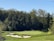 A lush green golf course featuring well-manicured grass and several sand bunkers is surrounded by dense, leafy trees. The clear blue sky enhances the picturesque landscape.