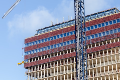 A partially constructed multi-story building with red brick-like panels and large glass windows. Steel beams are visible on the lower floors, indicating ongoing construction. Two cranes are positioned around the building, with one lifting machinery or materials. The sky is clear, contributing to good lighting conditions.