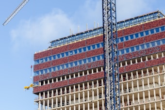 A partially constructed multi-story building with red brick-like panels and large glass windows. Steel beams are visible on the lower floors, indicating ongoing construction. Two cranes are positioned around the building, with one lifting machinery or materials. The sky is clear, contributing to good lighting conditions.