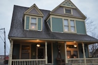 A two-story house with a steeply pitched roof and decorative shingles on the gables. The facade is painted in a muted teal color with cream trim, and the porch is lit with a warm glow from hanging lamps. There are decorative elements on the porch, including chairs and a wreath on the door.