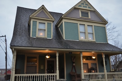 A two-story house with a steeply pitched roof and decorative shingles on the gables. The facade is painted in a muted teal color with cream trim, and the porch is lit with a warm glow from hanging lamps. There are decorative elements on the porch, including chairs and a wreath on the door.