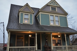 A two-story house with a steeply pitched roof and decorative shingles on the gables. The facade is painted in a muted teal color with cream trim, and the porch is lit with a warm glow from hanging lamps. There are decorative elements on the porch, including chairs and a wreath on the door.
