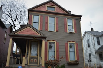 A two-story house with a symmetrical design, featuring brown siding and red shutters. The entrance includes small steps leading up to a porch with decorative columns. The house number is displayed above the door. Plants are visible in front of the house and inside window planters, and a tree without leaves stands next to the house.