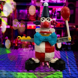 A colorful Lego clown figure is prominently displayed on a Lego baseplate. The clown is designed with a variety of bricks in bright colors like red, blue, yellow, and black. It features large eyes, a prominent red nose, and is holding a red lollipop-like object. The background is vibrant, filled with blurred lights and colors, creating a festive and playful atmosphere.