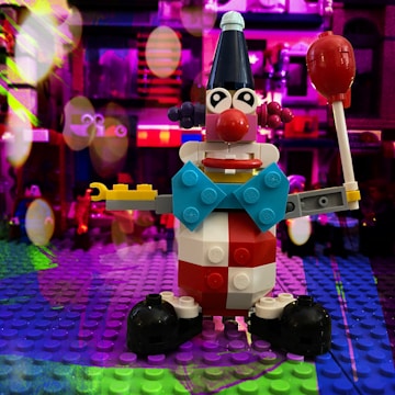 A colorful Lego clown figure is prominently displayed on a Lego baseplate. The clown is designed with a variety of bricks in bright colors like red, blue, yellow, and black. It features large eyes, a prominent red nose, and is holding a red lollipop-like object. The background is vibrant, filled with blurred lights and colors, creating a festive and playful atmosphere.
