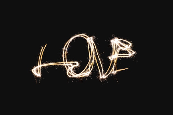 The word 'LOVE' is written in a sparkling light against a black background. The lettering appears to have been created with a sparkler or a similar light source, giving it a dynamic and festive appearance.