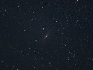 A starry night sky filled with countless stars. In the center, there is a faint nebulous object, likely a distant galaxy or nebula, with a subtle glow surrounded by the darkness of space.