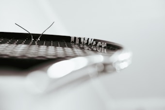 A close-up view of a tennis racket focusing on the strings. Two broken strings are visibly protruding, set against a blurred background. The image highlights the texture and pattern of the racket strings.