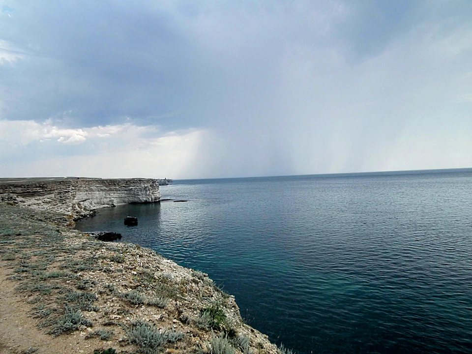 The image depicts a serene coastal scene with a rocky cliff overlooking a deep blue ocean. The sky above is filled with clouds, suggesting an overcast day. The perspective of the photo is from the shoreline, looking out towards the water. In the foreground, there are small rocks and shrubs scattered across the landscape.