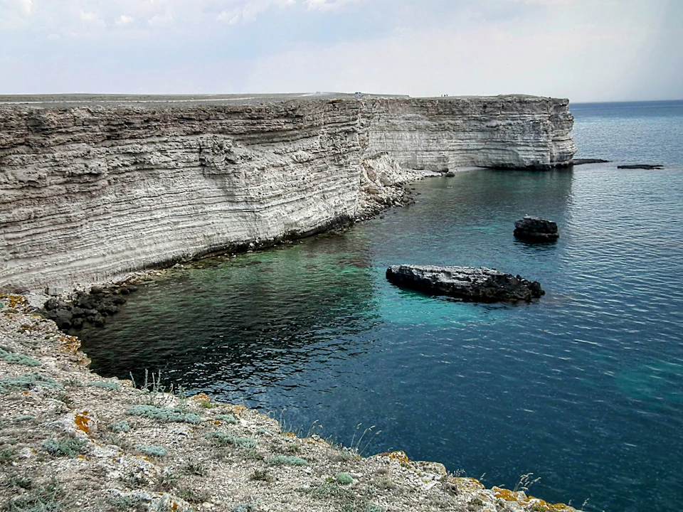The image depicts a rocky cliff overlooking a deep blue ocean. The cliff is composed of white and gray rock formations, with patches of green vegetation scattered across its surface. Two small rocks are visible on the right side of the cliff. The sky above is overcast, casting a soft light over the scene.