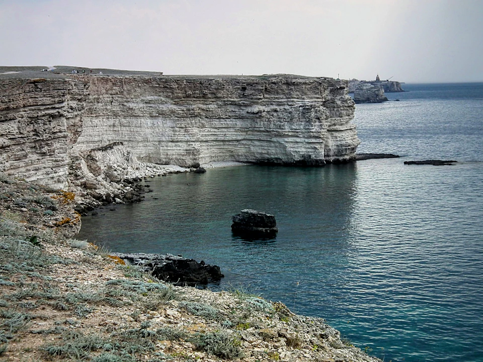 The image depicts a serene coastal landscape with a rocky cliff face on one side and a calm body of water on the other, both reflecting the overcast sky above. The cliff is covered in white rock formations that contrast with the darker hues of the surrounding environment. In the distance, beyond the cliff, there are buildings visible against the horizon line.
