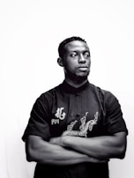 Man wearing a black shirt with deep blue embroidered details standing against a dark background.