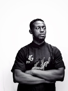 Man wearing a black shirt with deep blue embroidered details standing against a dark background.