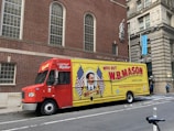 A delivery truck is parked on the street in front of a brick building with arched windows. The truck is primarily red and yellow with a large advertisement for W.B. Mason featuring a vintage-style portrait and American flags. The side of the truck prominently displays the company’s branding and slogan 'Who But W.B. Mason.' The surrounding area includes urban architecture and a sidewalk with pedestrians.