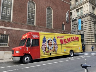 A delivery truck is parked on the street in front of a brick building with arched windows. The truck is primarily red and yellow with a large advertisement for W.B. Mason featuring a vintage-style portrait and American flags. The side of the truck prominently displays the company’s branding and slogan 'Who But W.B. Mason.' The surrounding area includes urban architecture and a sidewalk with pedestrians.