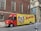 A delivery truck is parked on the street in front of a brick building with arched windows. The truck is primarily red and yellow with a large advertisement for W.B. Mason featuring a vintage-style portrait and American flags. The side of the truck prominently displays the company’s branding and slogan 'Who But W.B. Mason.' The surrounding area includes urban architecture and a sidewalk with pedestrians.