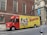A delivery truck is parked on the street in front of a brick building with arched windows. The truck is primarily red and yellow with a large advertisement for W.B. Mason featuring a vintage-style portrait and American flags. The side of the truck prominently displays the company’s branding and slogan 'Who But W.B. Mason.' The surrounding area includes urban architecture and a sidewalk with pedestrians.