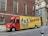 A delivery truck is parked on the street in front of a brick building with arched windows. The truck is primarily red and yellow with a large advertisement for W.B. Mason featuring a vintage-style portrait and American flags. The side of the truck prominently displays the company’s branding and slogan 'Who But W.B. Mason.' The surrounding area includes urban architecture and a sidewalk with pedestrians.