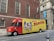 A delivery truck is parked on the street in front of a brick building with arched windows. The truck is primarily red and yellow with a large advertisement for W.B. Mason featuring a vintage-style portrait and American flags. The side of the truck prominently displays the company’s branding and slogan 'Who But W.B. Mason.' The surrounding area includes urban architecture and a sidewalk with pedestrians.