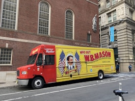 A delivery truck is parked on the street in front of a brick building with arched windows. The truck is primarily red and yellow with a large advertisement for W.B. Mason featuring a vintage-style portrait and American flags. The side of the truck prominently displays the company’s branding and slogan 'Who But W.B. Mason.' The surrounding area includes urban architecture and a sidewalk with pedestrians.