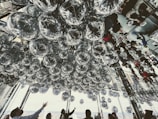 A mesmerizing ceiling installation made of numerous reflective silver spheres, creating an illusion of depth and distortion. The mirrored walls reflect the installation and the people below, adding complexity to the scene. People are gazing up, seemingly captivated by the artwork.