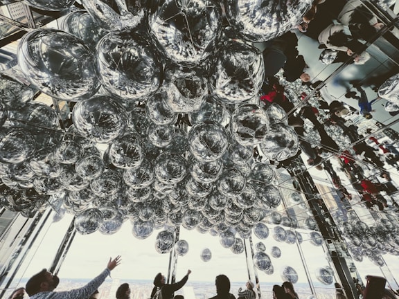 A mesmerizing ceiling installation made of numerous reflective silver spheres, creating an illusion of depth and distortion. The mirrored walls reflect the installation and the people below, adding complexity to the scene. People are gazing up, seemingly captivated by the artwork.