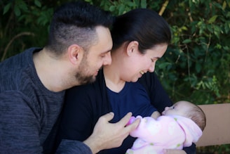 A couple lovingly interacts with a newborn baby, wrapped in a pink blanket, seated outdoors with greenery in the background. The woman is holding the baby and smiling, while the man looks on affectionately.