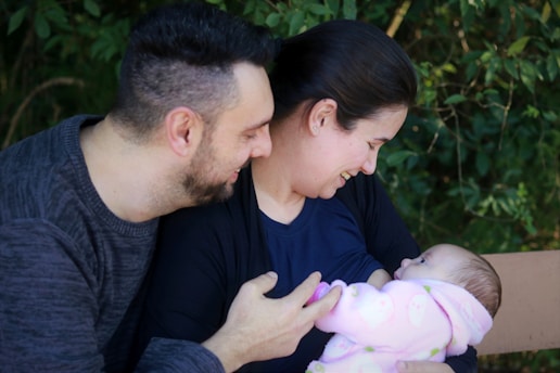 A couple lovingly interacts with a newborn baby, wrapped in a pink blanket, seated outdoors with greenery in the background. The woman is holding the baby and smiling, while the man looks on affectionately.
