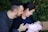 A couple lovingly interacts with a newborn baby, wrapped in a pink blanket, seated outdoors with greenery in the background. The woman is holding the baby and smiling, while the man looks on affectionately.