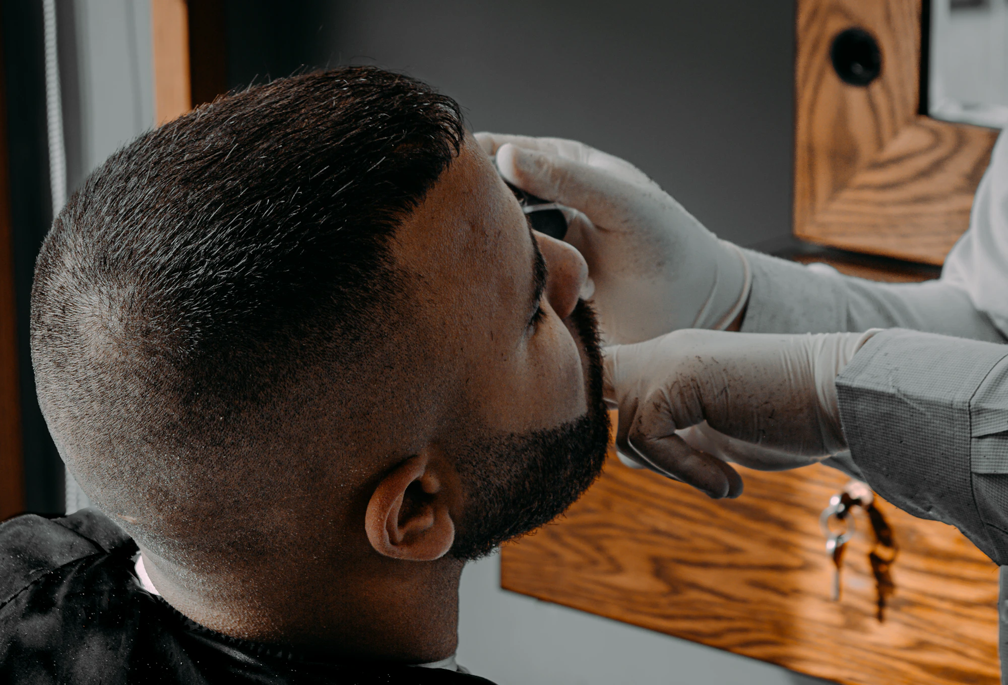 A man with a sharp skin fade and sculpted beard in profile while a barber works on his line up