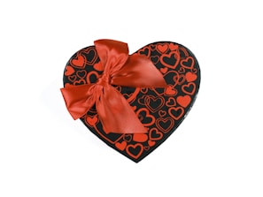 A heart-shaped gift box adorned with a large, bright red ribbon. The box is black with various red heart patterns covering its surface.