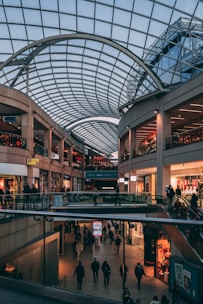A spacious indoor shopping mall with multiple levels, characterized by a large, arched glass ceiling that lets in natural light. Numerous shops line the sides, and people are walking and shopping in the mall.