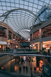 A spacious indoor shopping mall with multiple levels, characterized by a large, arched glass ceiling that lets in natural light. Numerous shops line the sides, and people are walking and shopping in the mall.