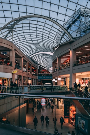 A spacious indoor shopping mall with multiple levels, characterized by a large, arched glass ceiling that lets in natural light. Numerous shops line the sides, and people are walking and shopping in the mall.