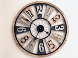 A large, round wall clock with a rustic design, featuring a mix of black, white, and wood-toned segments. Each segment holds a number made of metal, with a variety of fonts. The clock hands are thin and black, pointing at different angles.