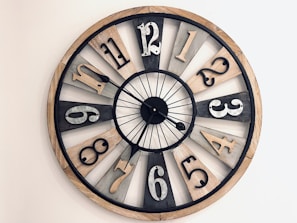 A large, round wall clock with a rustic design, featuring a mix of black, white, and wood-toned segments. Each segment holds a number made of metal, with a variety of fonts. The clock hands are thin and black, pointing at different angles.