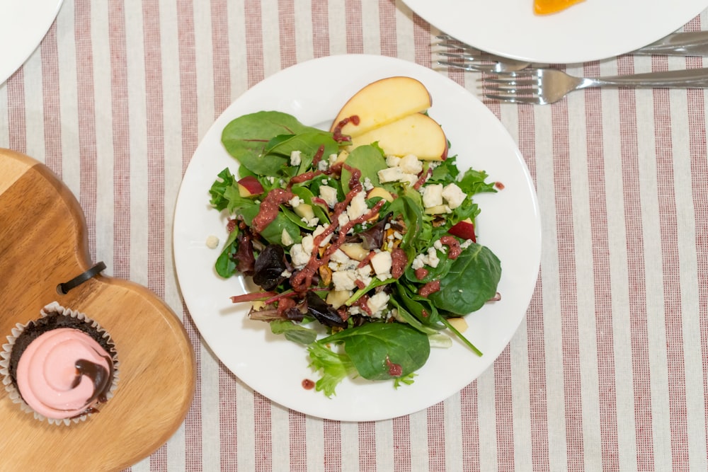 a white plate topped with a salad next to a cupcake