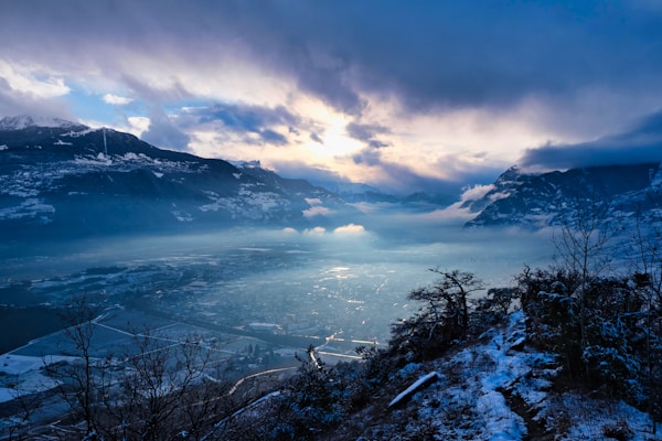 A breathtaking mountain landscape with snow-covered peaks and a dramatic sky, partially illuminated by the setting or rising sun. A valley below is shrouded in mist, creating a serene and mystical atmosphere. Sparse snow-dusted vegetation is visible in the foreground.