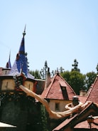 A whimsical building with tall, pointed roofs covered in blue and red tiles, resembling a fairytale castle. Long, golden hair drapes from one of the windows, hinting at a storybook theme. Surrounding the structure are trees under a clear blue sky.